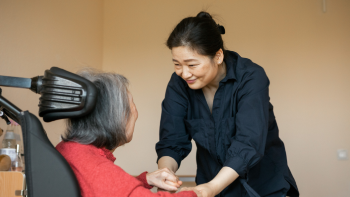 Eine Frau von hinten sitzt im Rollstuhl. Sie hat graue Haare. Ihr Kopf wird von einer Kopfstütze gehalten. Ihr gegenüber steht eine asiatische Frau mittleren Alters. Sie beugt sich zur Frau im Rollstuhl, lächelt ihr zu und hält ihre beiden Hände. 