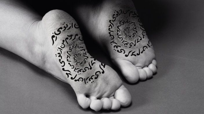 The black and white photo shows a pair of outstretched female feet. We look at the soles of the feet, which are inscribed with Persian letters