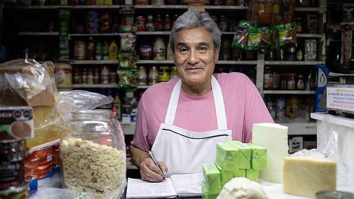 A man stands behind the crowded sales counter of a store and looks into the camera with a smile. The shelves behind him are full of cans and packages