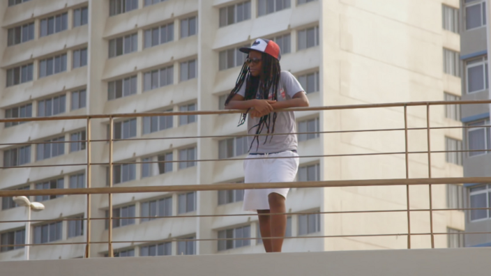 Suthu Magiwane steht an einem Geländer. Unter ihrem roten Basecap kommen ihre langen dunklen Haare hervor. Sie trägt Sonnenbrille. Im Hintergrund steht ein Hochhaus.