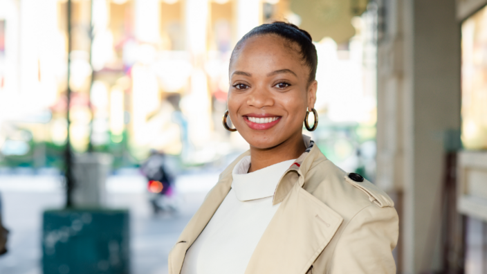 A portrait of a young black woman. She wears a trench coat, large round earrings and smiles into the camera.