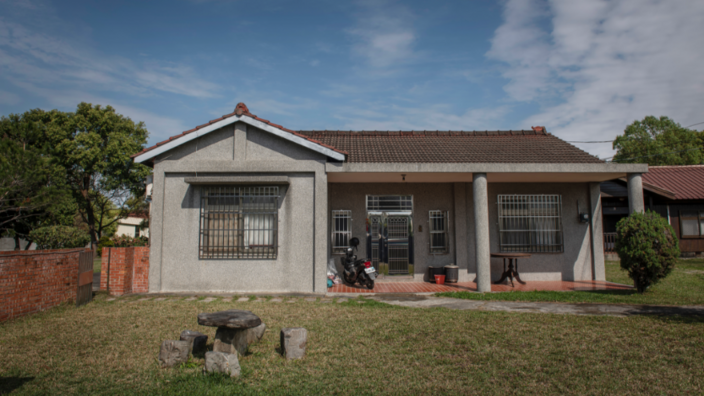 A gray concrete house stands on a plot of land. A simple wooden seating area is on the lawn in front of the house. Another table and a two-wheeler are on the terrace.