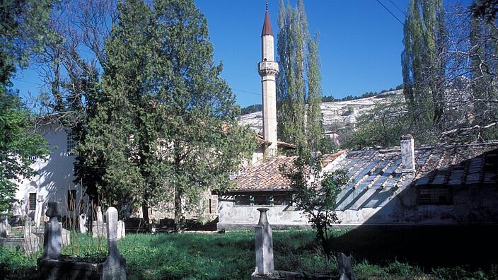 On a plot with a lawn there are several monuments made of stone. Behind the lawn you can see a narrow tower, two houses and several trees. 