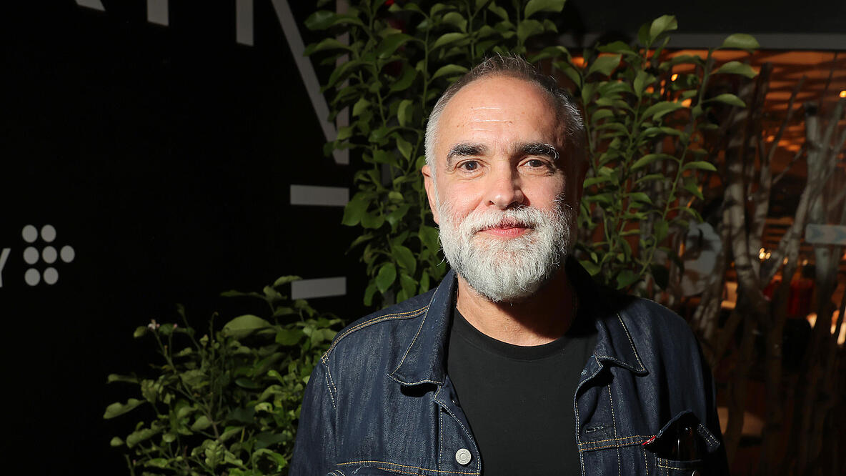 A man with a grey beard in a dark room, lit from the side. He is wearing a denim jacket and a black T-shirt, his hands in his trouser pockets. He looks friendly into the camera