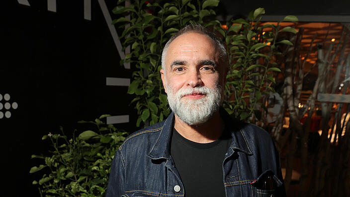 A man with a grey beard in a dark room, lit from the side. He is wearing a denim jacket and a black T-shirt, his hands in his trouser pockets. He looks friendly into the camera