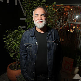 A man with a grey beard in a dark room, lit from the side. He is wearing a denim jacket and a black T-shirt, his hands in his trouser pockets. He looks friendly into the camera