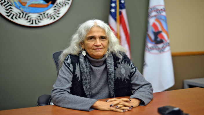 A slightly older woman sits at a desk with folded hands, behind her two flags are vaguely visible. She has long white hair, wears silver earrings and a gray sweater with an overcoat. She looks directly into the camera.