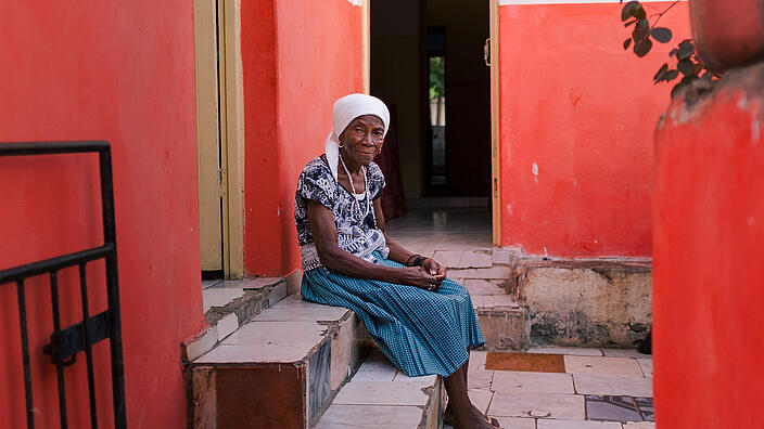 Eine ältere schwarze Frau sitzt vor ihrem Haus auf den Treppenstufen. 