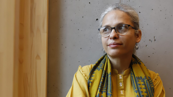 A woman in a mustard yellow shirt with a patterned scarf is leaning against a railing in front of a concrete wall next to a window. She wears glasses and her long grey hair is tied back. She looks thoughtfully out of the window to the side