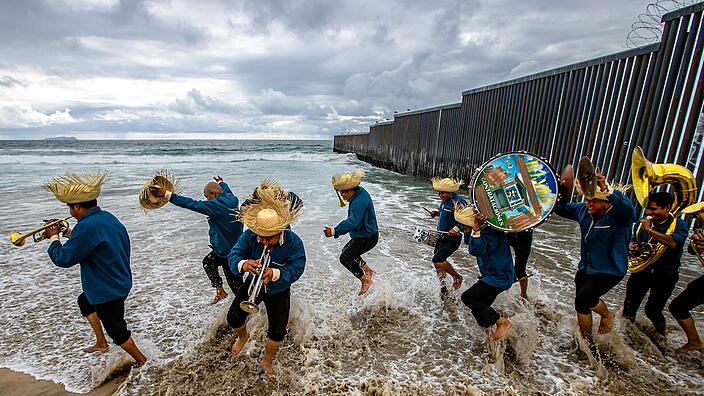 On the beach in Tijuana, a Mexican band dances with their feet in the water while playing their instruments. The men are wearing a dark blue shirt, black trousers and straw hats. They play a drum, a timpani and brass instruments. The border wall between Mexico and the USA can be seen in the background, protruding into the water