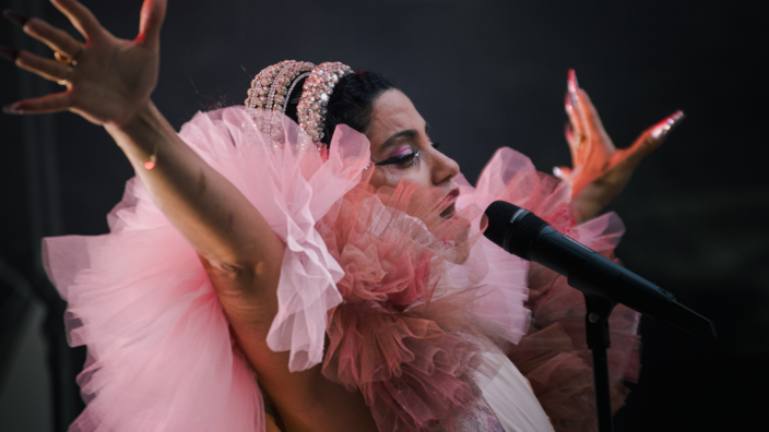 Die tunesische Sängerin Emel Mathlouthi steht am Mikrofon vor dunklem Hintergrund. Sie trägt ein extravagantes Kleid mit vielen rosa Rüschen und glitzerndem Haarschmuck. Beim Singen breitet sie dramatisch die Arme aus. 